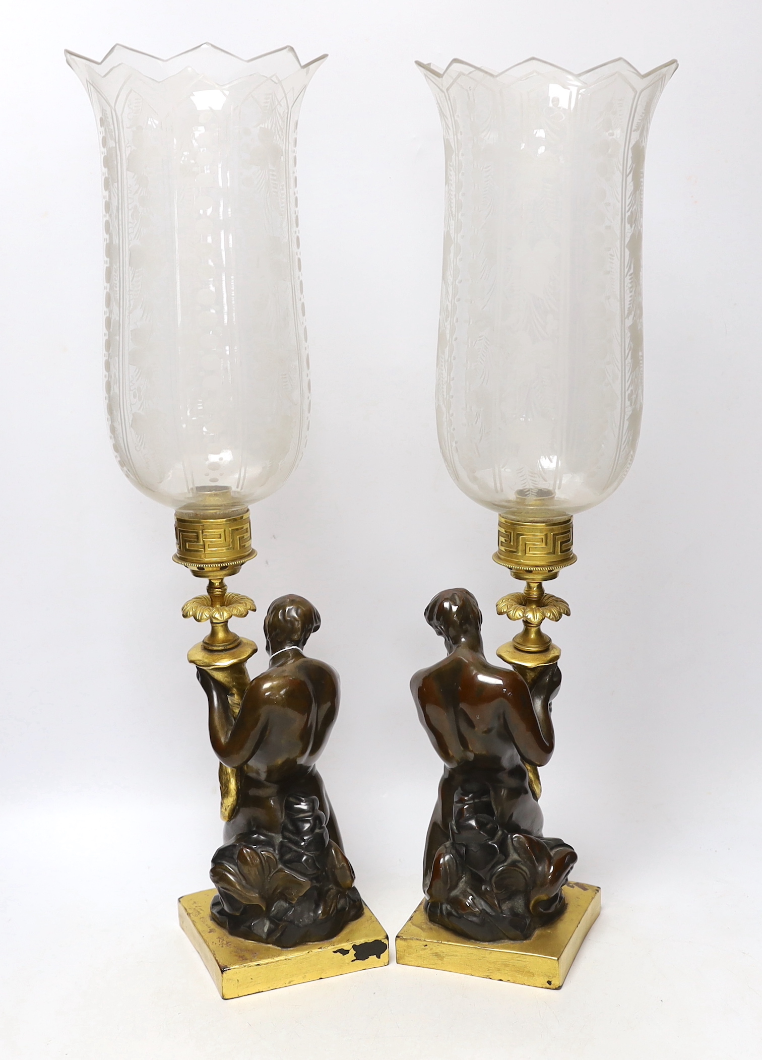 A pair of 19th century bronzed stoneware candlesticks, after a design by John Flaxman, each modelled as a triton holding a cornucopia, supporting a gilt brass sconce and engraved glass hurricane shade, 54cm high